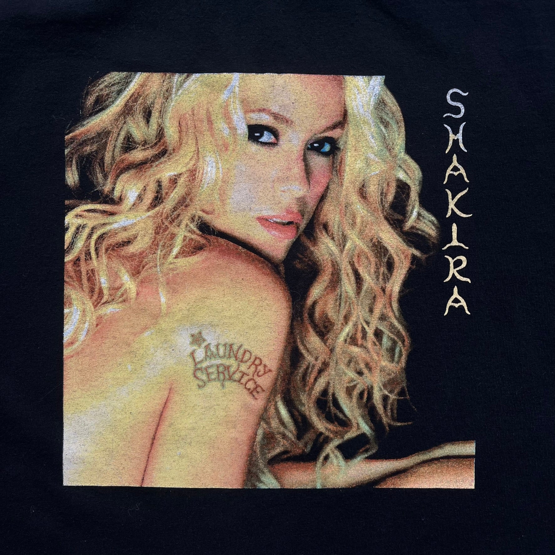 Shakira 2002 “Tour de la Mangosta” Concert Tee is a black cotton T-shirt from the 2002–2003 tour merchandise range. The front features a square photo of Shakira with yellow “Shakira” text alongside the image. The back includes a large “Tour de la Mangosta” print with a serpent logo and the tour dates 2002–2003, followed by a list of concert locations.
