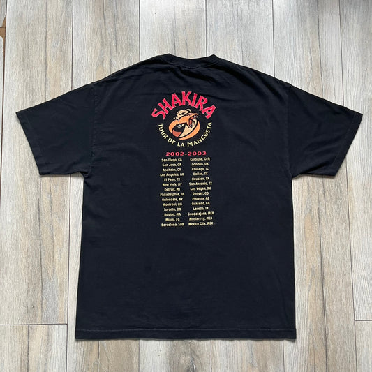 Shakira 2002 “Tour de la Mangosta” Concert Tee is a black cotton T-shirt from the 2002–2003 tour merchandise range. The front features a square photo of Shakira with yellow “Shakira” text alongside the image. The back includes a large “Tour de la Mangosta” print with a serpent logo and the tour dates 2002–2003, followed by a list of concert locations.
