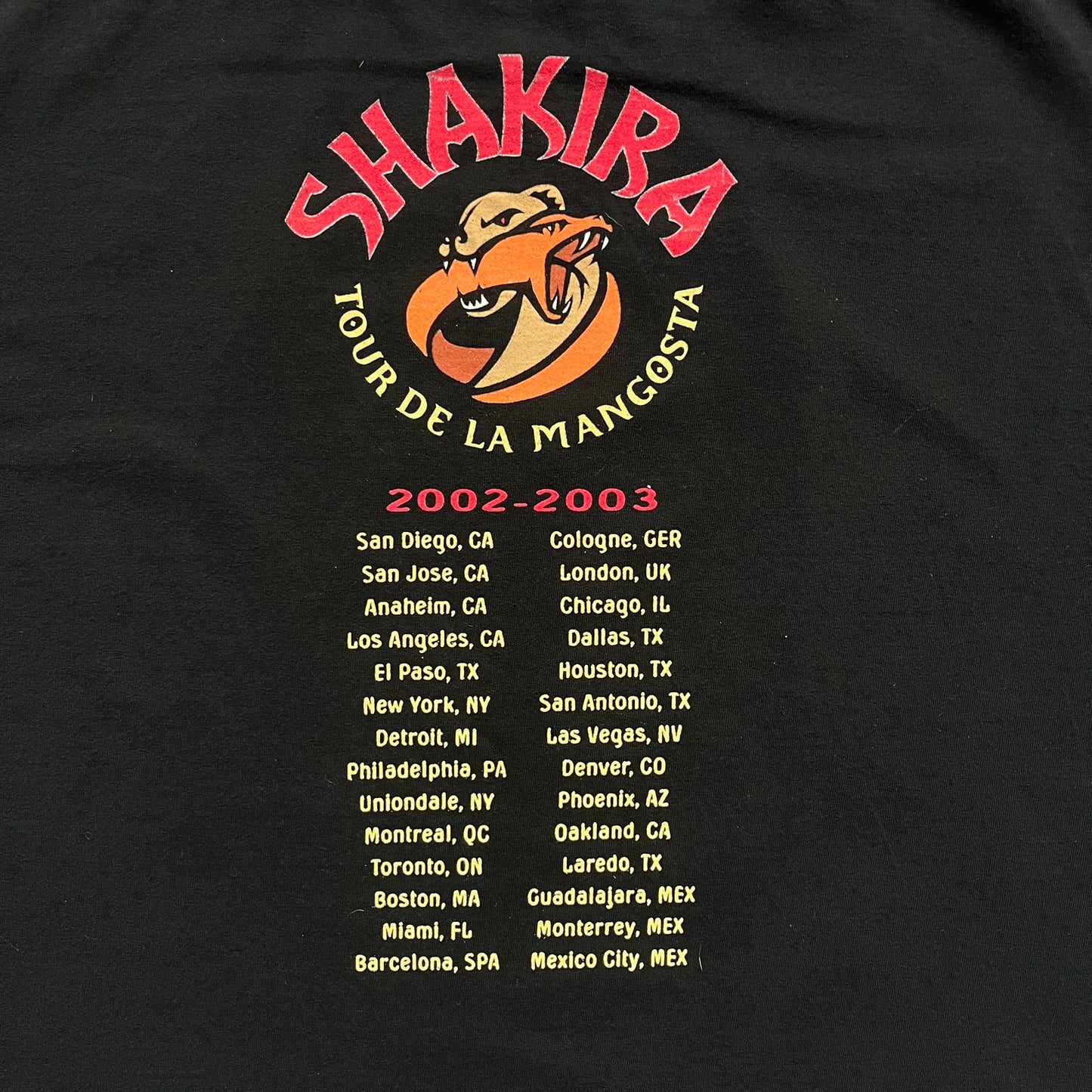 Shakira 2002 “Tour de la Mangosta” Concert Tee is a black cotton T-shirt from the 2002–2003 tour merchandise range. The front features a square photo of Shakira with yellow “Shakira” text alongside the image. The back includes a large “Tour de la Mangosta” print with a serpent logo and the tour dates 2002–2003, followed by a list of concert locations.
