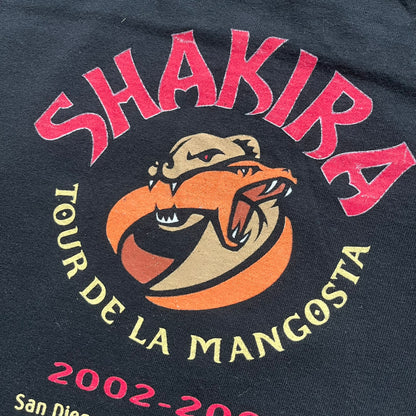 Shakira 2002 “Tour de la Mangosta” Concert Tee is a black cotton T-shirt from the 2002–2003 tour merchandise range. The front features a square photo of Shakira with yellow “Shakira” text alongside the image. The back includes a large “Tour de la Mangosta” print with a serpent logo and the tour dates 2002–2003, followed by a list of concert locations.
