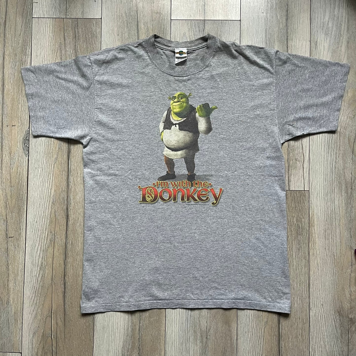 Shrek 2003 “I’m with the Donkey” Gray Tee is a gray T-shirt made from 90% cotton and 10% polyester. The front features Shrek pointing to the side with “I’m with the Donkey” text in a medieval font, and a small DreamWorks Shrek 2003 licensing print below. The neck tag shows Shrek and Universal Studios branding.