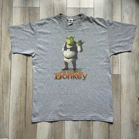 Shrek 2003 “I’m with the Donkey” Gray Tee is a gray T-shirt made from 90% cotton and 10% polyester. The front features Shrek pointing to the side with “I’m with the Donkey” text in a medieval font, and a small DreamWorks Shrek 2003 licensing print below. The neck tag shows Shrek and Universal Studios branding.