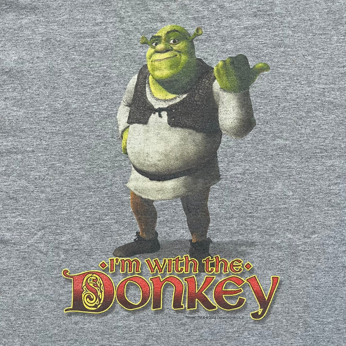 Shrek 2003 “I’m with the Donkey” Gray Tee is a gray T-shirt made from 90% cotton and 10% polyester. The front features Shrek pointing to the side with “I’m with the Donkey” text in a medieval font, and a small DreamWorks Shrek 2003 licensing print below. The neck tag shows Shrek and Universal Studios branding.