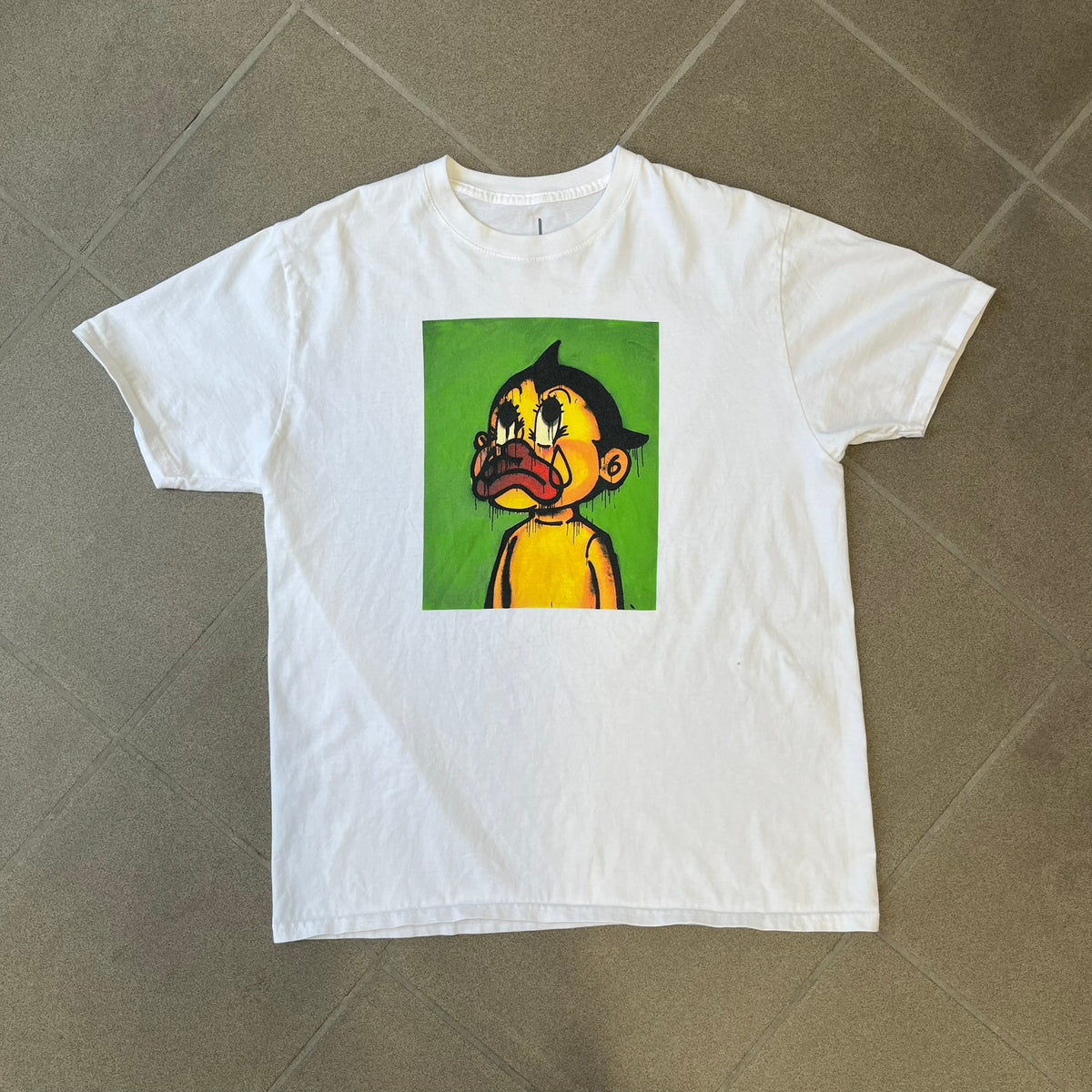 Slawn 2025 White Astroboy Tee is a white T-shirt featuring a rectangular graphic of a yellow Astroboy-inspired figure with a sad Slawn-style face and black hair, set against a green background. It carries a Slawn neck tag.