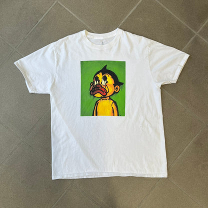 Slawn 2025 White Astroboy Tee is a white T-shirt featuring a rectangular graphic of a yellow Astroboy-inspired figure with a sad Slawn-style face and black hair, set against a green background. It carries a Slawn neck tag.