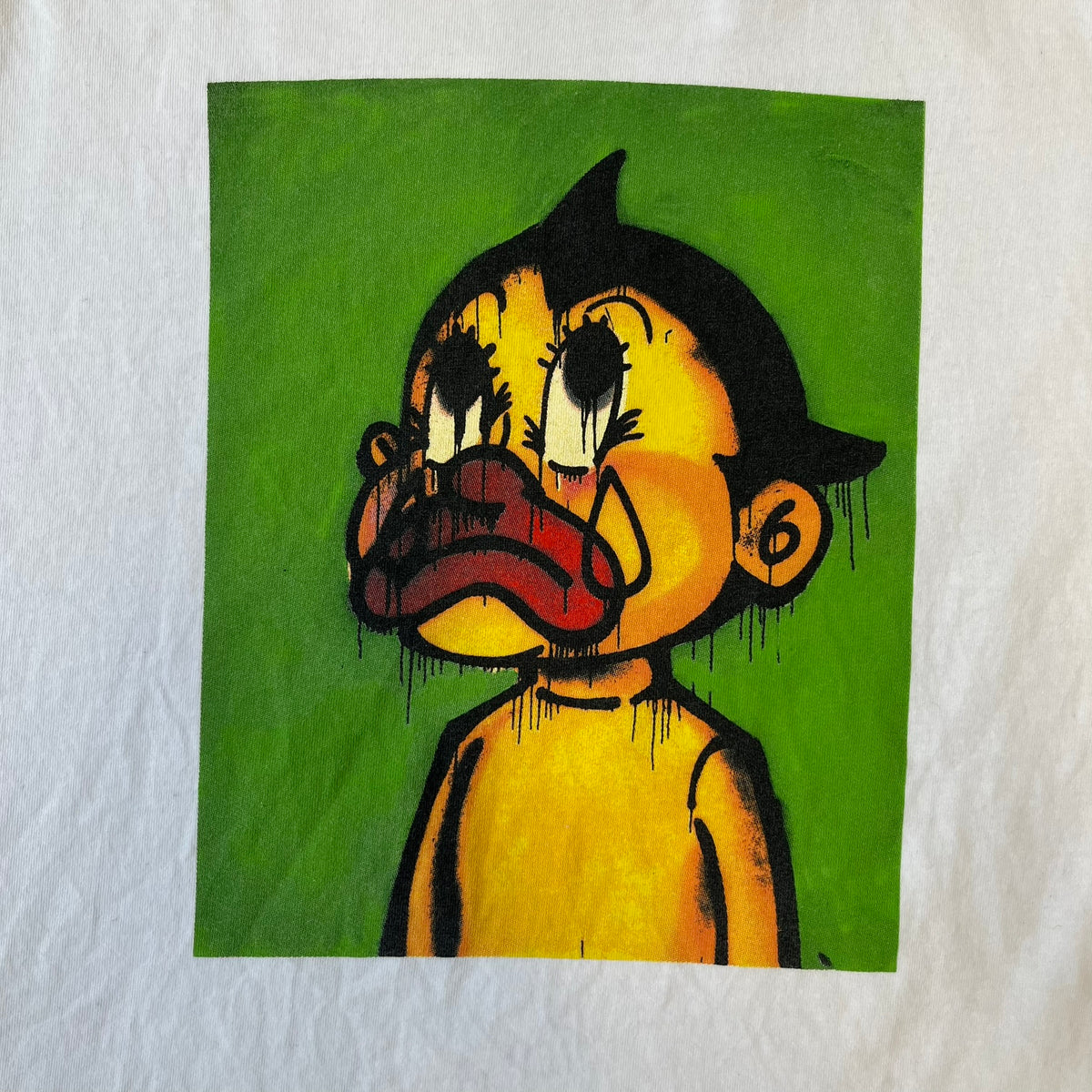 Slawn 2025 White Astroboy Tee is a white T-shirt featuring a rectangular graphic of a yellow Astroboy-inspired figure with a sad Slawn-style face and black hair, set against a green background. It carries a Slawn neck tag.