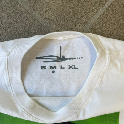 Slawn 2025 White Astroboy Tee is a white T-shirt featuring a rectangular graphic of a yellow Astroboy-inspired figure with a sad Slawn-style face and black hair, set against a green background. It carries a Slawn neck tag.