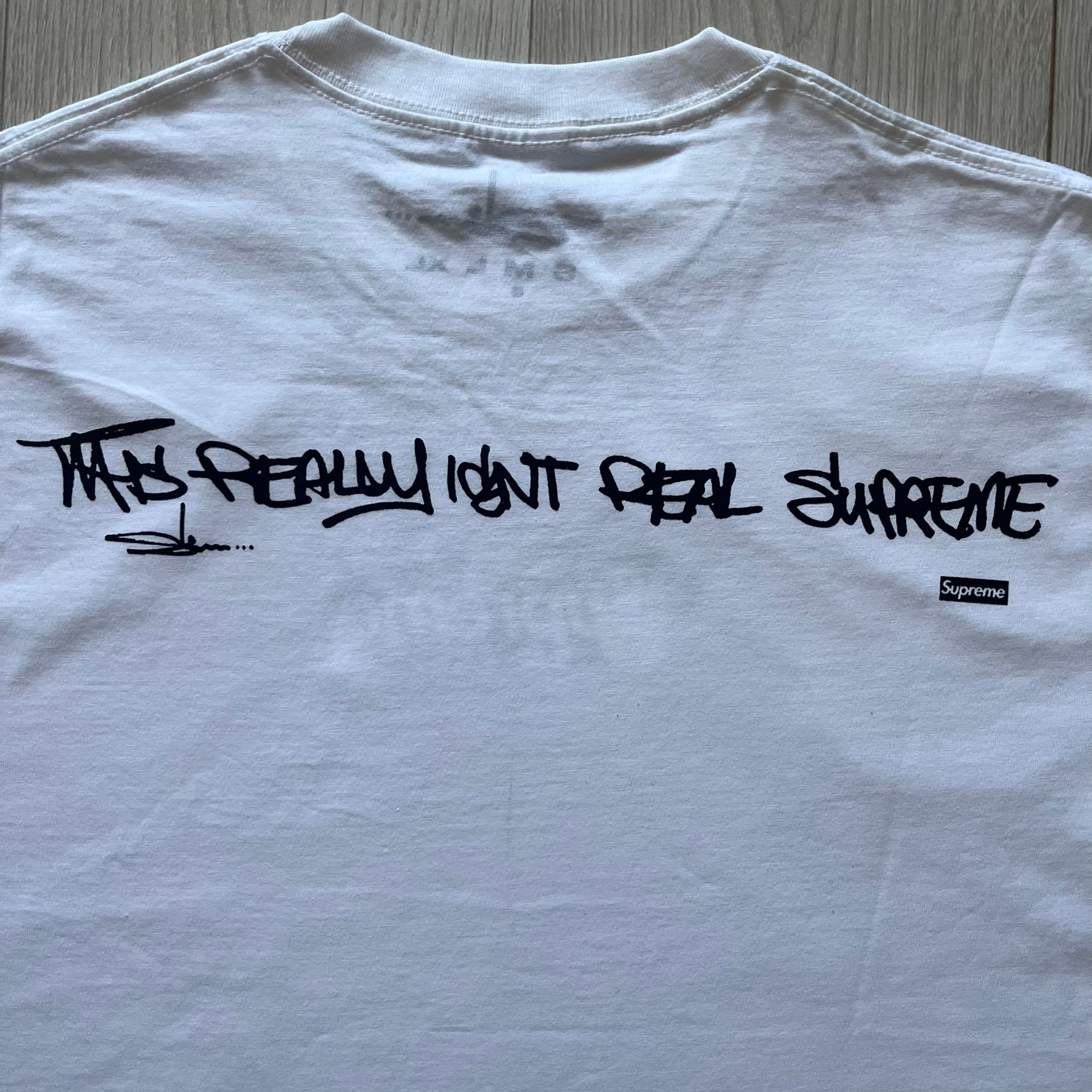 White Slawn Batpreme tee featuring a Batman-inspired graphic with iconic twist by Slawn and Supreme text overlay. Printed neck tag shows Slawn’s signature, while the back reads “This really isn’t real Supreme” in hand-drawn style with signature and logo. A striking, collectible streetwear piece.