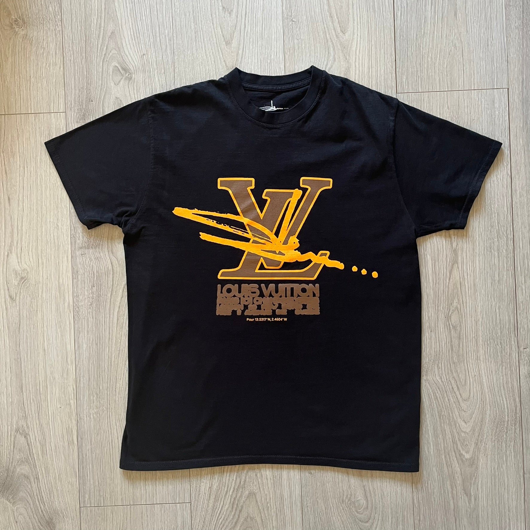 Black t-shirt with gold and brown graphic design on a wooden floor
