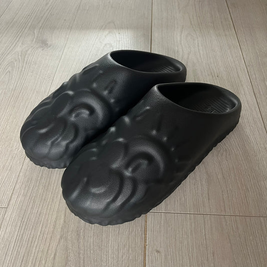 Slawn Shapes Black Chompaz Sliders in the black colorway, designed by artist Olaolu Slawn in collaboration with Shape Shoes. These closed-toe rubber sliders feature Slawn’s signature face design as the body of the shoe. They include a custom tread pattern that leaves a unique footprint. Packaged in a cardboard box with a special plastic bag covered in Slawn’s signature face graphics.