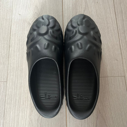 Slawn Shapes Black Chompaz Sliders in the black colorway, designed by artist Olaolu Slawn in collaboration with Shape Shoes. These closed-toe rubber sliders feature Slawn’s signature face design as the body of the shoe. They include a custom tread pattern that leaves a unique footprint. Packaged in a cardboard box with a special plastic bag covered in Slawn’s signature face graphics.