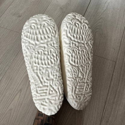 Slawn Shapes Snow Stompaz Sliders in the white colorway, created in collaboration with artist Olaolu Slawn and Shape Shoes. These closed-toe rubber sliders feature Slawn’s signature cartoonish face design forming the body of the shoe and the playful tagline “F*ck Crusty Toes.” The tread is specially designed to leave a unique footprint. They come packaged in a cardboard box filled with snow-like wool and include a special silver sticker.