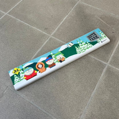 South Park 1998 Keyboard Wrist Rest is a thick foam wrist rest featuring the iconic bus stop scene with Cartman, Kenny, Stan, and Kyle kicking Ike. The design includes the wooden South Park sign and a small 1998 Comedy Central licensing mark, with a black base.