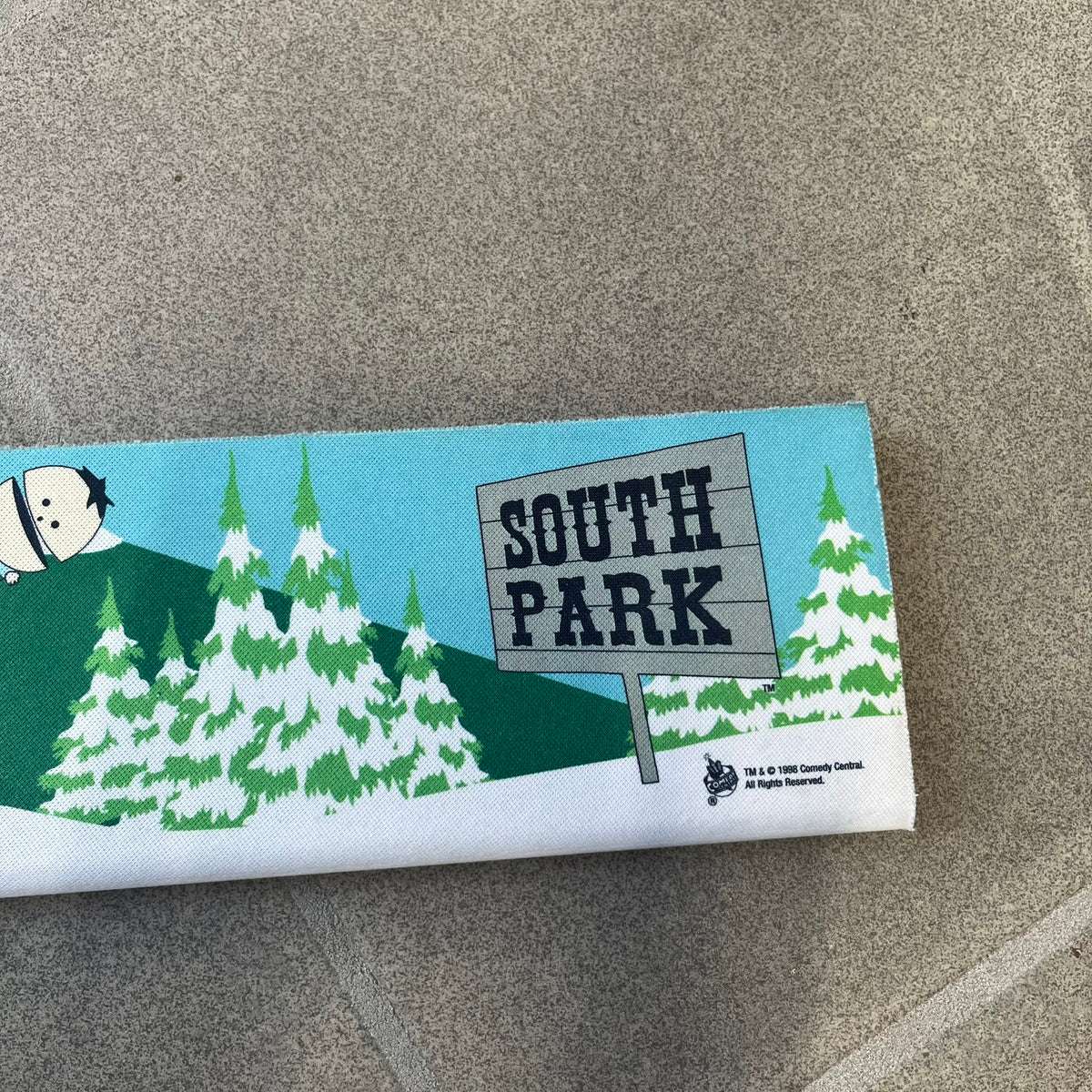 South Park 1998 Keyboard Wrist Rest is a thick foam wrist rest featuring the iconic bus stop scene with Cartman, Kenny, Stan, and Kyle kicking Ike. The design includes the wooden South Park sign and a small 1998 Comedy Central licensing mark, with a black base.