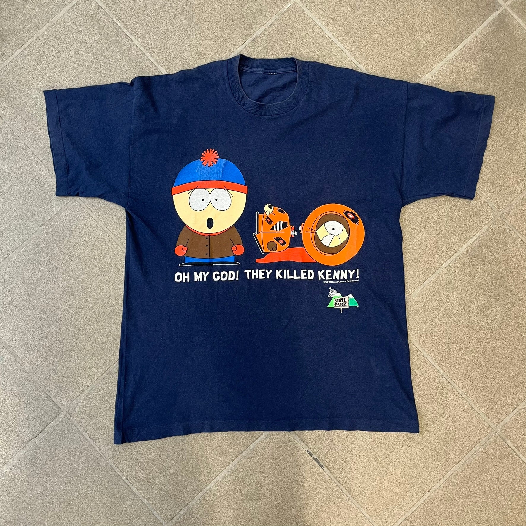 South Park 1998 “They Killed Kenny” Navy Tee features a a front graphic of Stan standing with a surprised expression next to a fallen Kenny. Below the characters is white “Oh My God! They Killed Kenny!” text, with a small South Park logo and 1998 licensing detail printed underneath on the right side. Made from cotton.