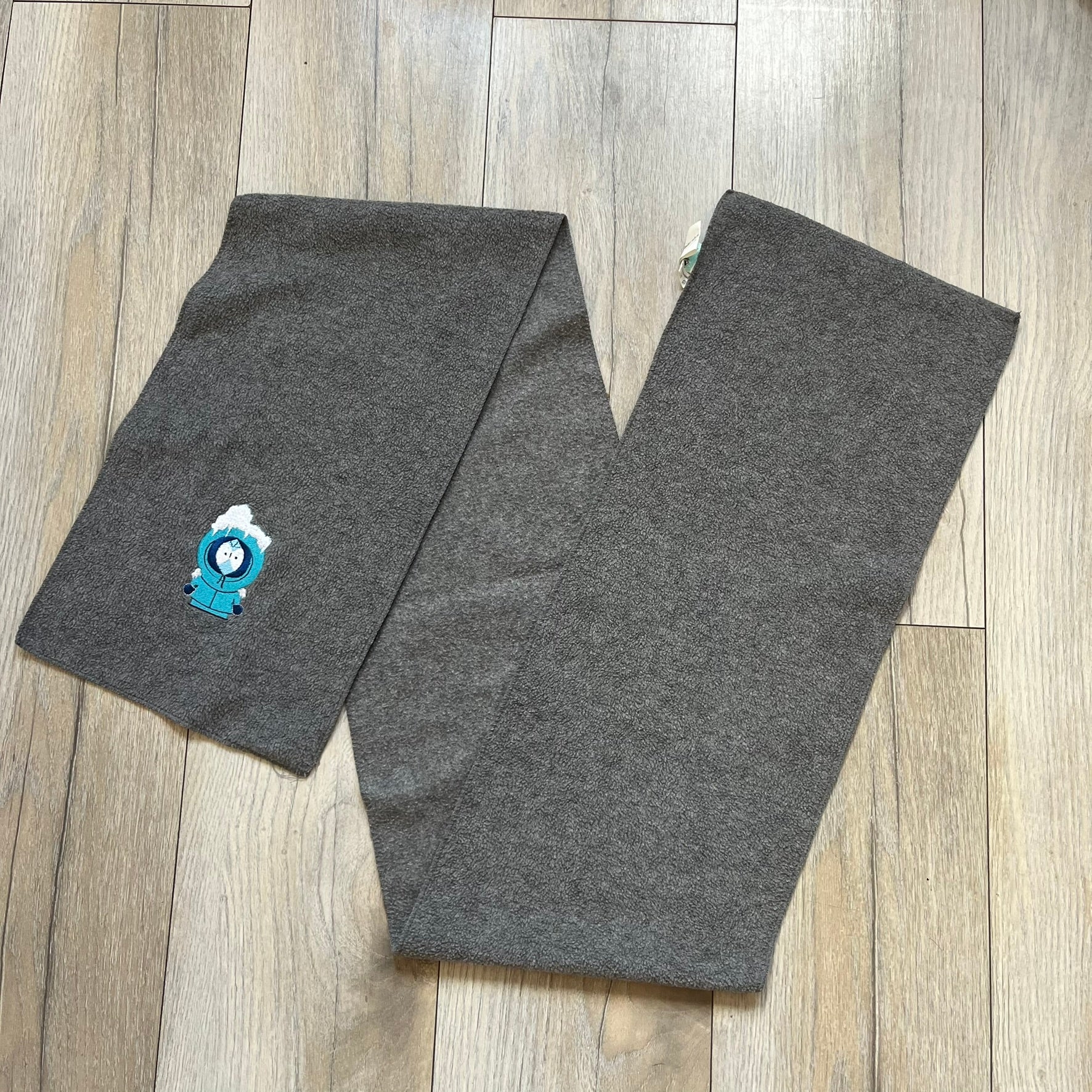 South Park 2000 Frozen Kenny Scarf is a grey scarf featuring a graphic of Kenny rendered in a frozen state in blue tones. The character is shown with ice and snow effects covering the head area. The scarf includes a South Park Comedy Central tag and 2000 licensing details.
