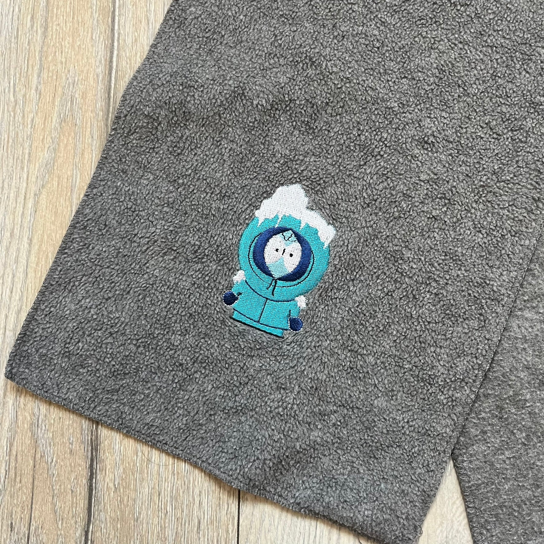 South Park 2000 Frozen Kenny Scarf is a grey scarf featuring a graphic of Kenny rendered in a frozen state in blue tones. The character is shown with ice and snow effects covering the head area. The scarf includes a South Park Comedy Central tag and 2000 licensing details.
