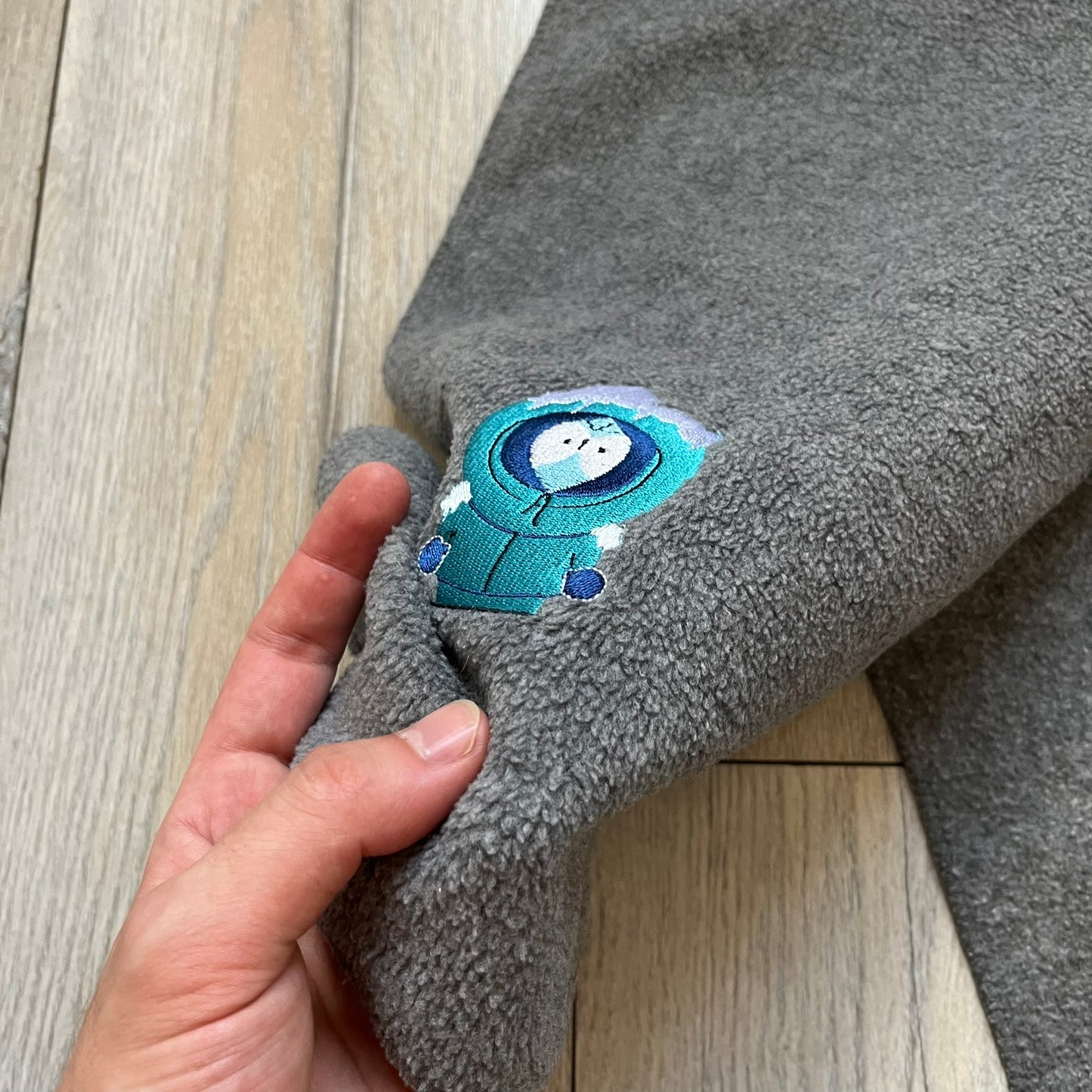 South Park 2000 Frozen Kenny Scarf is a grey scarf featuring a graphic of Kenny rendered in a frozen state in blue tones. The character is shown with ice and snow effects covering the head area. The scarf includes a South Park Comedy Central tag and 2000 licensing details.
