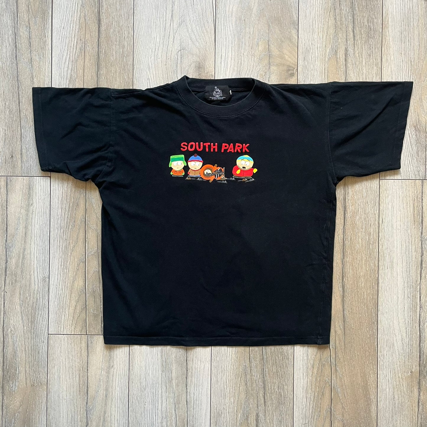 South Park 2000 Kenny’s Rats Black Tee is a black short-sleeve T-shirt with a Comedy Central 2000 licensed neck tag. The front features red “South Park” text above a graphic of Kenny, Stan, and Cartman standing while Kenny is depicted on the ground being eaten by rats. The shirt has a simple boxy fit with a front print design.