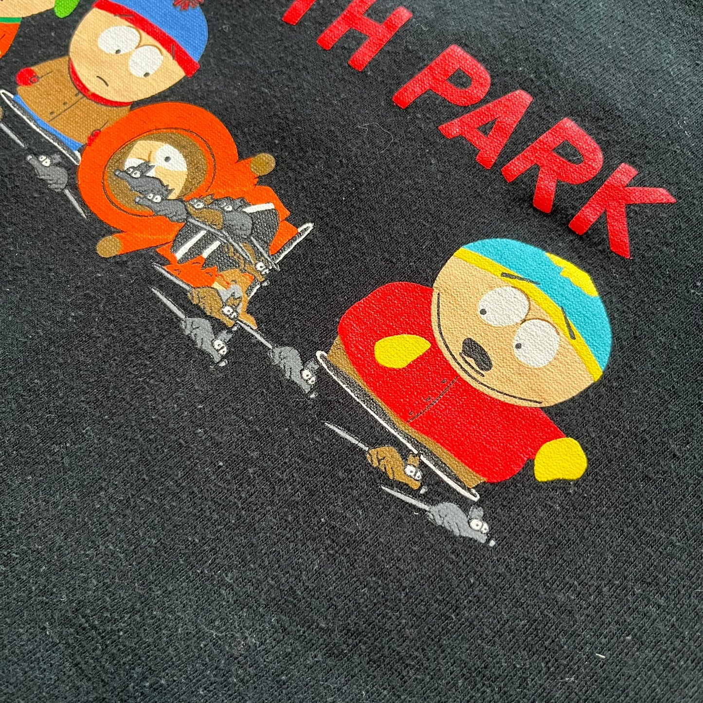 South Park 2000 Kenny’s Rats Black Tee is a black short-sleeve T-shirt with a Comedy Central 2000 licensed neck tag. The front features red “South Park” text above a graphic of Kenny, Stan, and Cartman standing while Kenny is depicted on the ground being eaten by rats. The shirt has a simple boxy fit with a front print design.