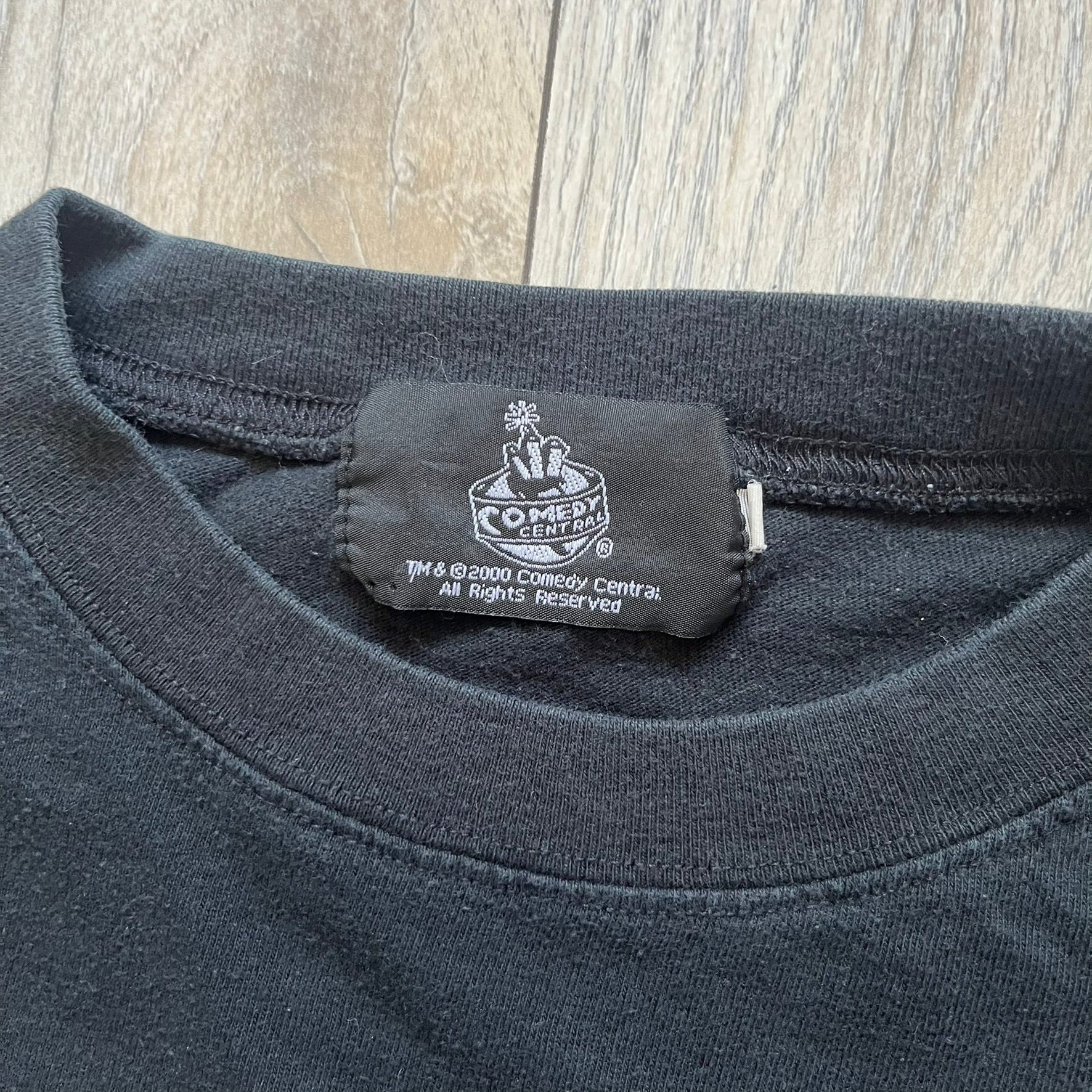 South Park 2000 Kenny’s Rats Black Tee is a black short-sleeve T-shirt with a Comedy Central 2000 licensed neck tag. The front features red “South Park” text above a graphic of Kenny, Stan, and Cartman standing while Kenny is depicted on the ground being eaten by rats. The shirt has a simple boxy fit with a front print design.