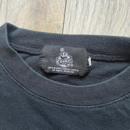 South Park 2000 Kenny’s Rats Black Tee is a black short-sleeve T-shirt with a Comedy Central 2000 licensed neck tag. The front features red “South Park” text above a graphic of Kenny, Stan, and Cartman standing while Kenny is depicted on the ground being eaten by rats. The shirt has a simple boxy fit with a front print design.