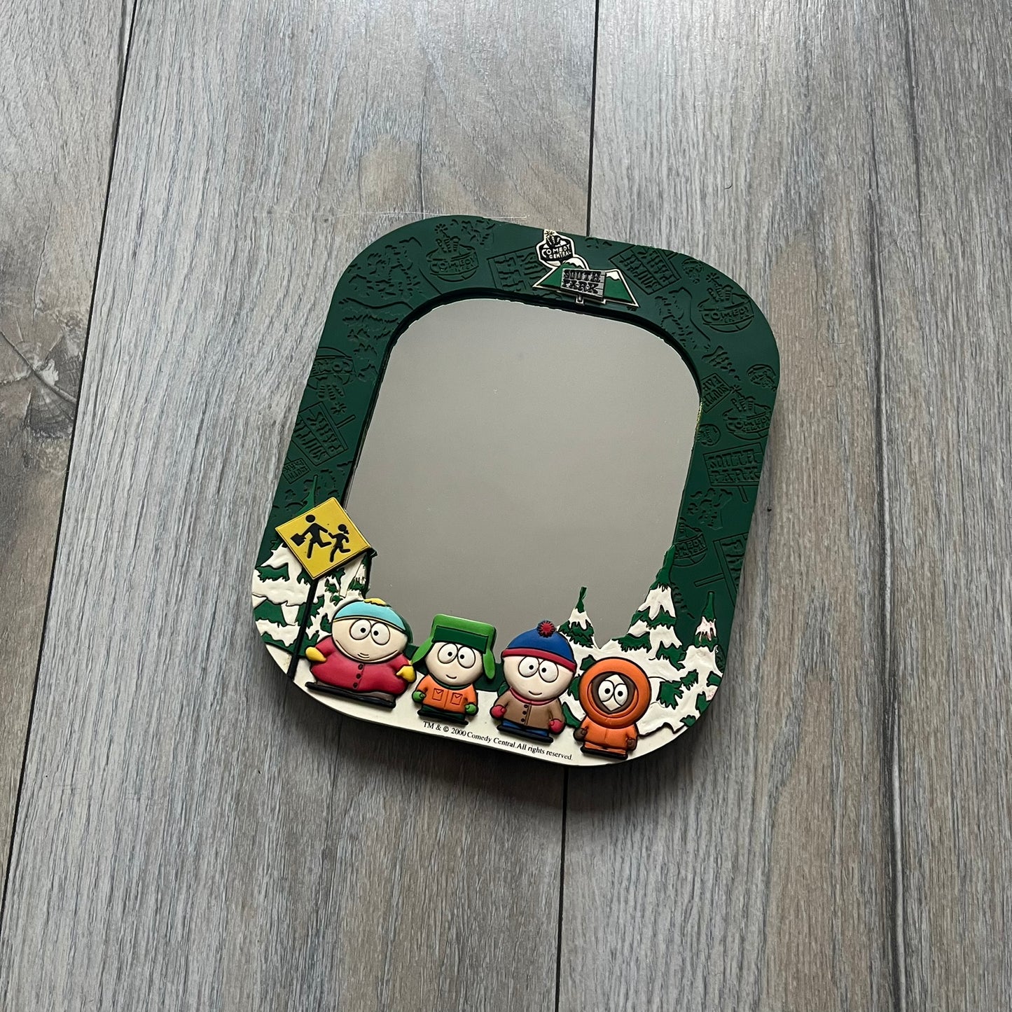 Small tabletop mirror with a molded rubber frame featuring all-over cutout logos for South Park and Comedy Central, along with leaf and tree motifs. The top is finished with a raised 3D South Park logo, while the bottom features 3D rubber figures of Cartman, Kenny, Kyle, and Stan standing at their bus stop. Marked with a 2000 Comedy Central copyright at the bottom, with a black backing and integrated stand.