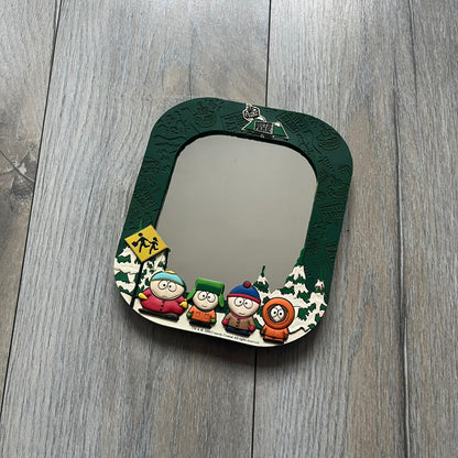 Small tabletop mirror with a molded rubber frame featuring all-over cutout logos for South Park and Comedy Central, along with leaf and tree motifs. The top is finished with a raised 3D South Park logo, while the bottom features 3D rubber figures of Cartman, Kenny, Kyle, and Stan standing at their bus stop. Marked with a 2000 Comedy Central copyright at the bottom, with a black backing and integrated stand.