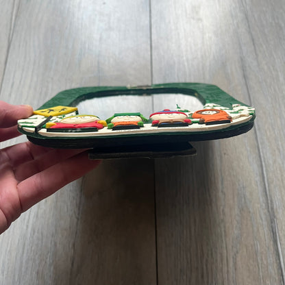 Small tabletop mirror with a molded rubber frame featuring all-over cutout logos for South Park and Comedy Central, along with leaf and tree motifs. The top is finished with a raised 3D South Park logo, while the bottom features 3D rubber figures of Cartman, Kenny, Kyle, and Stan standing at their bus stop. Marked with a 2000 Comedy Central copyright at the bottom, with a black backing and integrated stand.