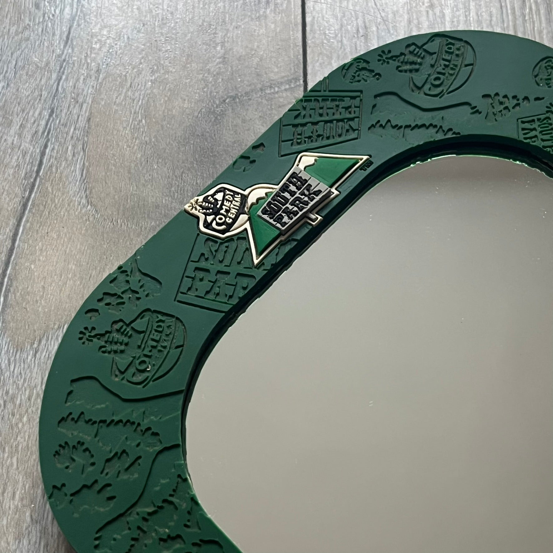 Small tabletop mirror with a molded rubber frame featuring all-over cutout logos for South Park and Comedy Central, along with leaf and tree motifs. The top is finished with a raised 3D South Park logo, while the bottom features 3D rubber figures of Cartman, Kenny, Kyle, and Stan standing at their bus stop. Marked with a 2000 Comedy Central copyright at the bottom, with a black backing and integrated stand.