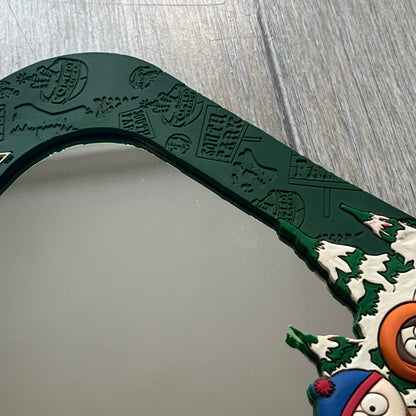 Small tabletop mirror with a molded rubber frame featuring all-over cutout logos for South Park and Comedy Central, along with leaf and tree motifs. The top is finished with a raised 3D South Park logo, while the bottom features 3D rubber figures of Cartman, Kenny, Kyle, and Stan standing at their bus stop. Marked with a 2000 Comedy Central copyright at the bottom, with a black backing and integrated stand.