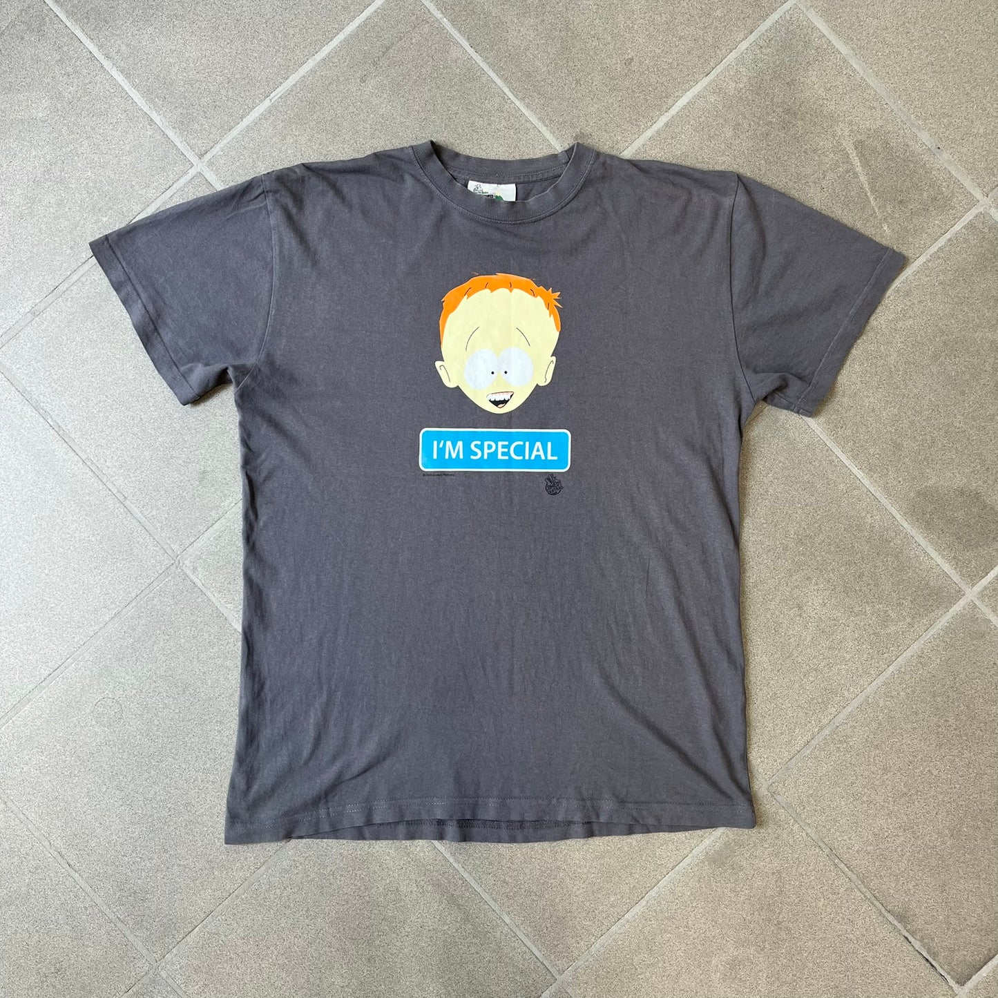 South Park 2008 Timmy “I’m Special” Tee features a grey T-shirt with a front graphic of Timmy’s face above a blue bar with white “I’m Special” text. Small 2008 licensing and Comedy Central details are printed underneath. The back features a blue and white wheelchair-accessibility sign graphic. Includes a licensed South Park neck tag.
