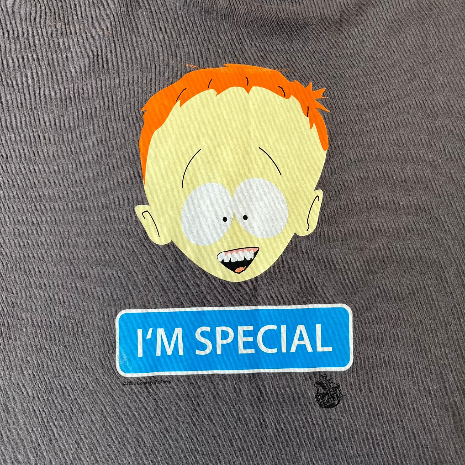 South Park 2008 Timmy “I’m Special” Tee features a grey T-shirt with a front graphic of Timmy’s face above a blue bar with white “I’m Special” text. Small 2008 licensing and Comedy Central details are printed underneath. The back features a blue and white wheelchair-accessibility sign graphic. Includes a licensed South Park neck tag.
