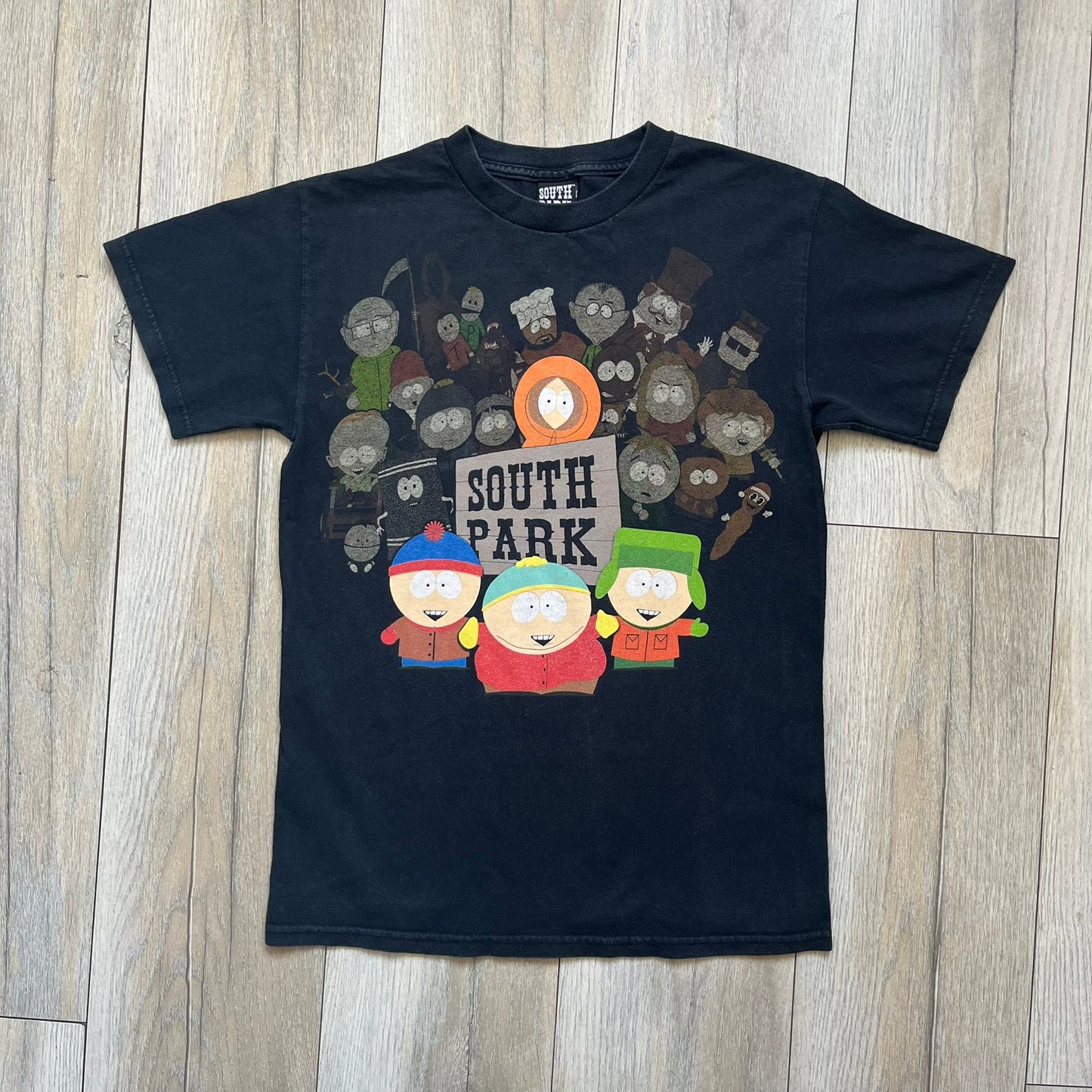 South Park 2009 Cast Tee is a black T-shirt featuring a large front graphic of Stan, Kyle, Cartman, and Kenny standing beside a wooden South Park sign. Behind them is a background of approximately twenty additional South Park characters printed in washed tones. The shirt includes a South Park neck tag with 2009 licensing.