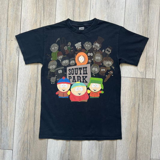 South Park 2009 Cast Tee is a black T-shirt featuring a large front graphic of Stan, Kyle, Cartman, and Kenny standing beside a wooden South Park sign. Behind them is a background of approximately twenty additional South Park characters printed in washed tones. The shirt includes a South Park neck tag with 2009 licensing.