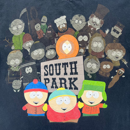 South Park 2009 Cast Tee is a black T-shirt featuring a large front graphic of Stan, Kyle, Cartman, and Kenny standing beside a wooden South Park sign. Behind them is a background of approximately twenty additional South Park characters printed in washed tones. The shirt includes a South Park neck tag with 2009 licensing.