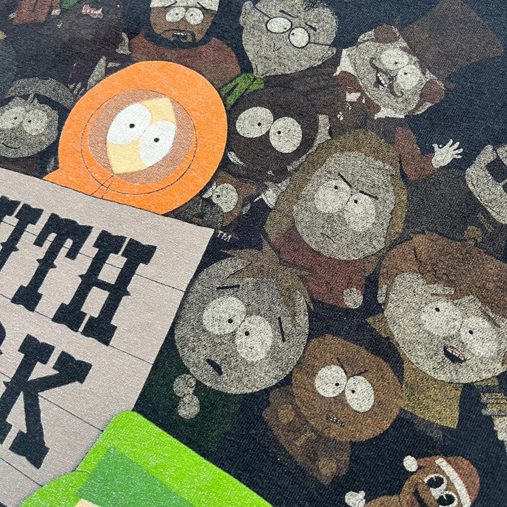 South Park 2009 Cast Tee is a black T-shirt featuring a large front graphic of Stan, Kyle, Cartman, and Kenny standing beside a wooden South Park sign. Behind them is a background of approximately twenty additional South Park characters printed in washed tones. The shirt includes a South Park neck tag with 2009 licensing.