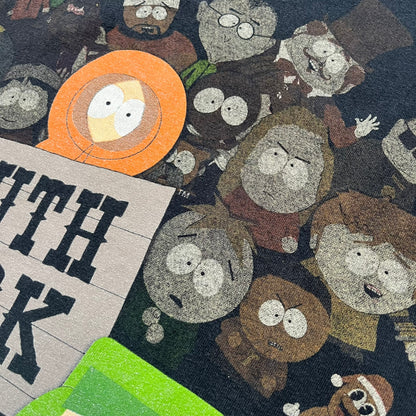 South Park 2009 Cast Tee is a black T-shirt featuring a large front graphic of Stan, Kyle, Cartman, and Kenny standing beside a wooden South Park sign. Behind them is a background of approximately twenty additional South Park characters printed in washed tones. The shirt includes a South Park neck tag with 2009 licensing.