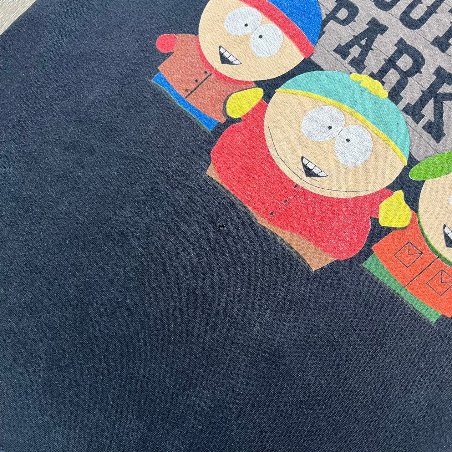 South Park 2009 Cast Tee is a black T-shirt featuring a large front graphic of Stan, Kyle, Cartman, and Kenny standing beside a wooden South Park sign. Behind them is a background of approximately twenty additional South Park characters printed in washed tones. The shirt includes a South Park neck tag with 2009 licensing.