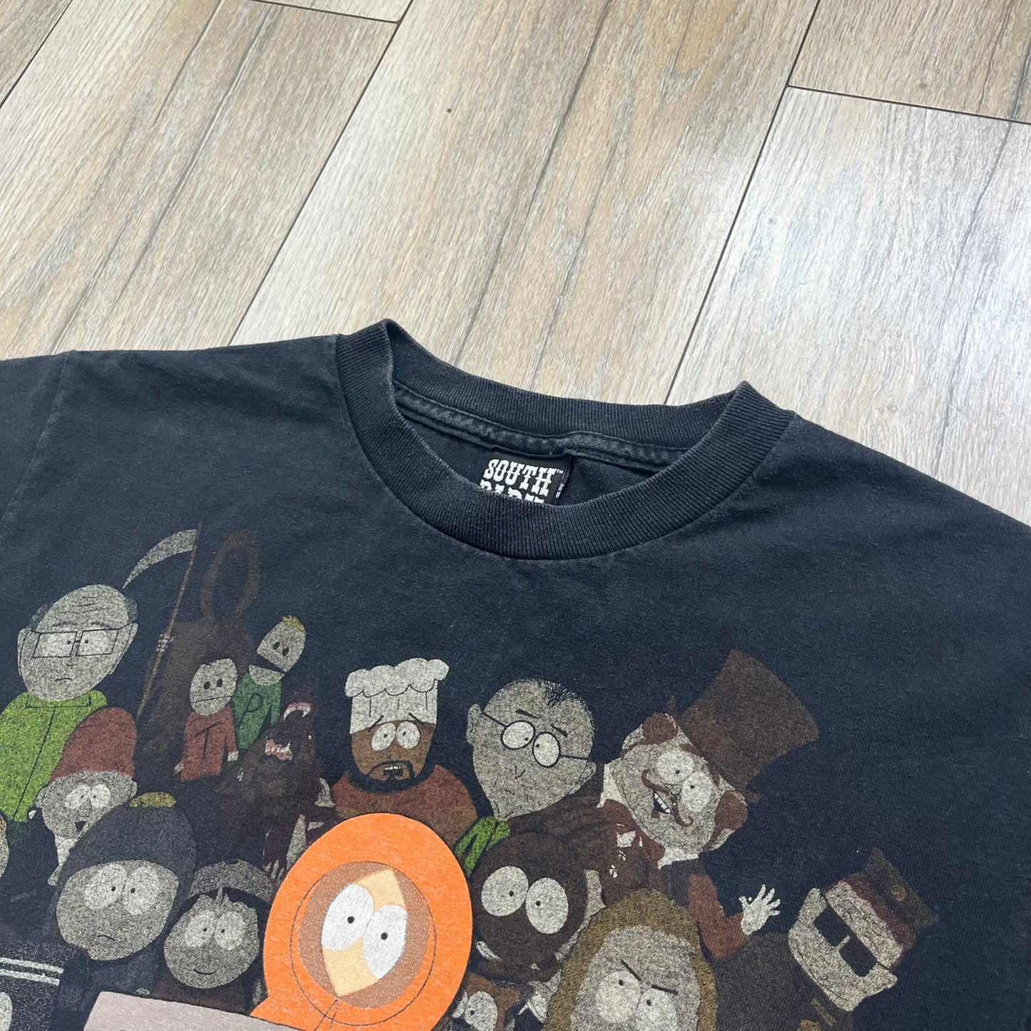 South Park 2009 Cast Tee is a black T-shirt featuring a large front graphic of Stan, Kyle, Cartman, and Kenny standing beside a wooden South Park sign. Behind them is a background of approximately twenty additional South Park characters printed in washed tones. The shirt includes a South Park neck tag with 2009 licensing.