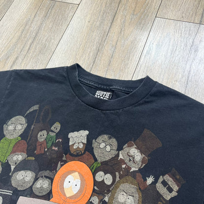 South Park 2009 Cast Tee is a black T-shirt featuring a large front graphic of Stan, Kyle, Cartman, and Kenny standing beside a wooden South Park sign. Behind them is a background of approximately twenty additional South Park characters printed in washed tones. The shirt includes a South Park neck tag with 2009 licensing.
