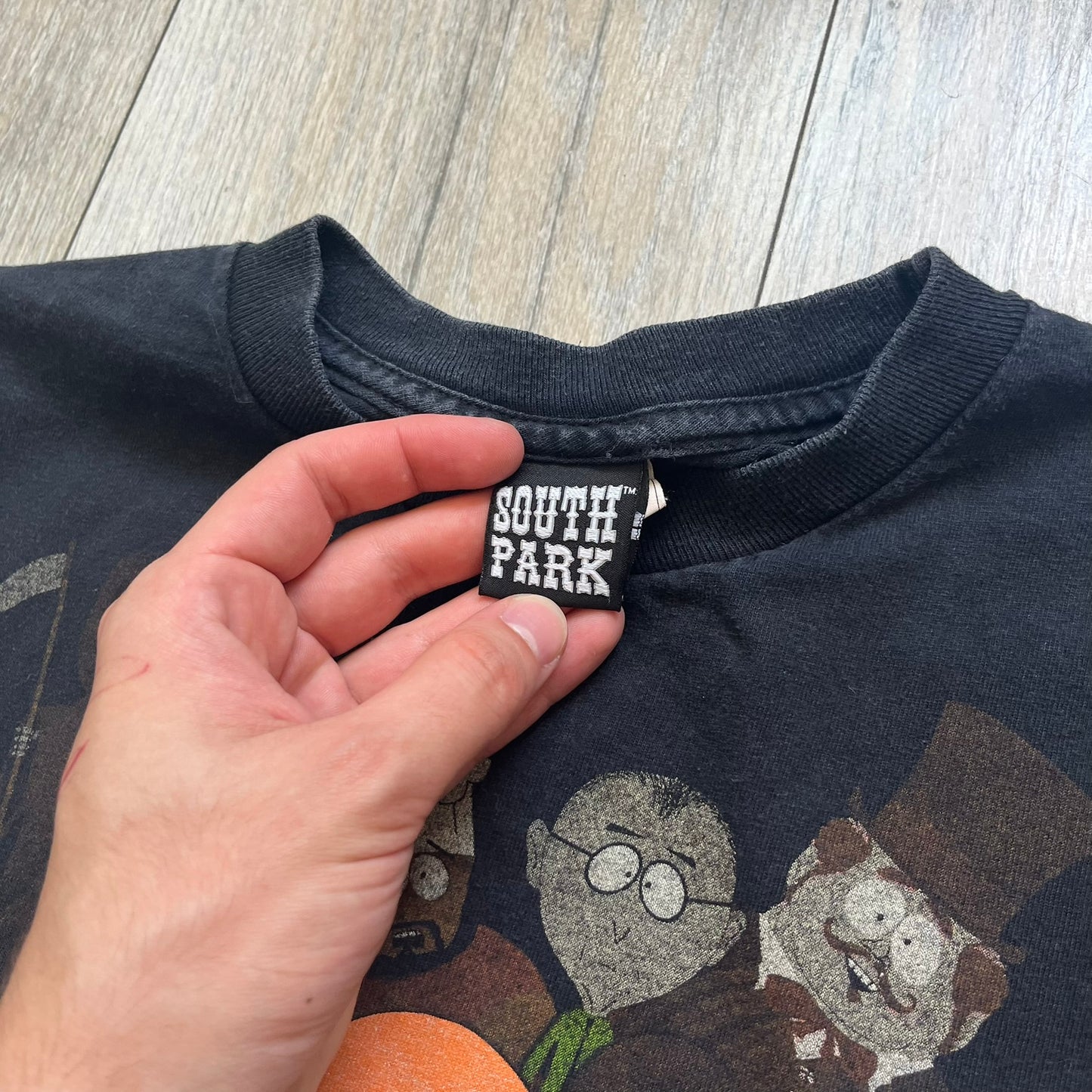 South Park 2009 Cast Tee is a black T-shirt featuring a large front graphic of Stan, Kyle, Cartman, and Kenny standing beside a wooden South Park sign. Behind them is a background of approximately twenty additional South Park characters printed in washed tones. The shirt includes a South Park neck tag with 2009 licensing.