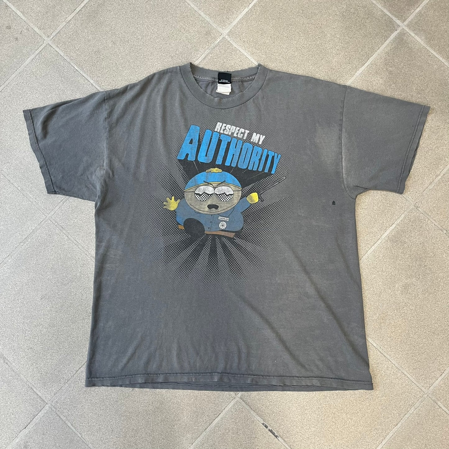 South Park 2009 “Respect My Authority” Gray Tee features a grey T-shirt with a front graphic of Cartman wearing a police uniform and running across a dotted cartoon-style background. Above the graphic is white and blue “Respect My Authority” text. The shirt has a South Park 2009 licensed neck tag and a heavily worn, distressed condition.
