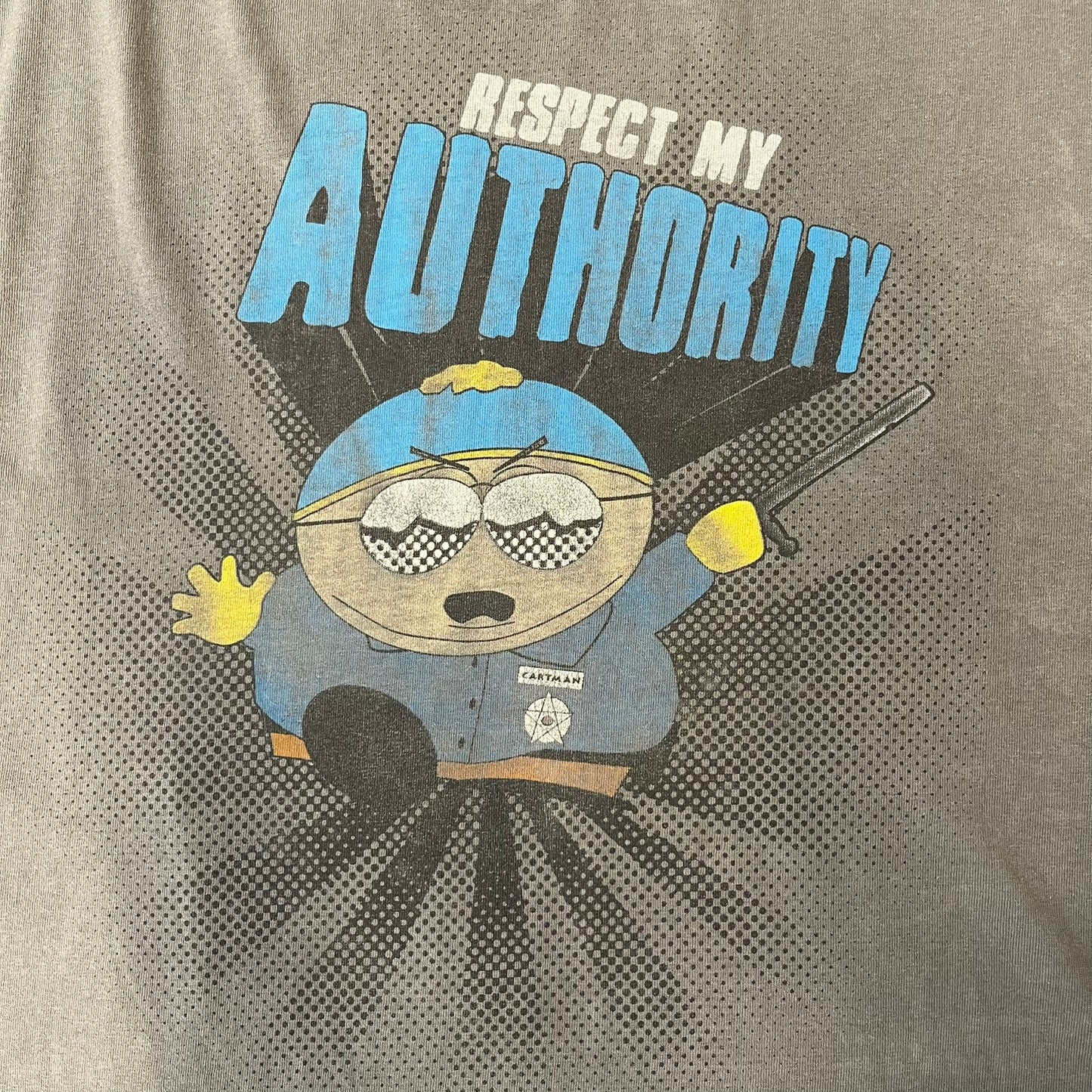 South Park 2009 “Respect My Authority” Gray Tee features a grey T-shirt with a front graphic of Cartman wearing a police uniform and running across a dotted cartoon-style background. Above the graphic is white and blue “Respect My Authority” text. The shirt has a South Park 2009 licensed neck tag and a heavily worn, distressed condition.
