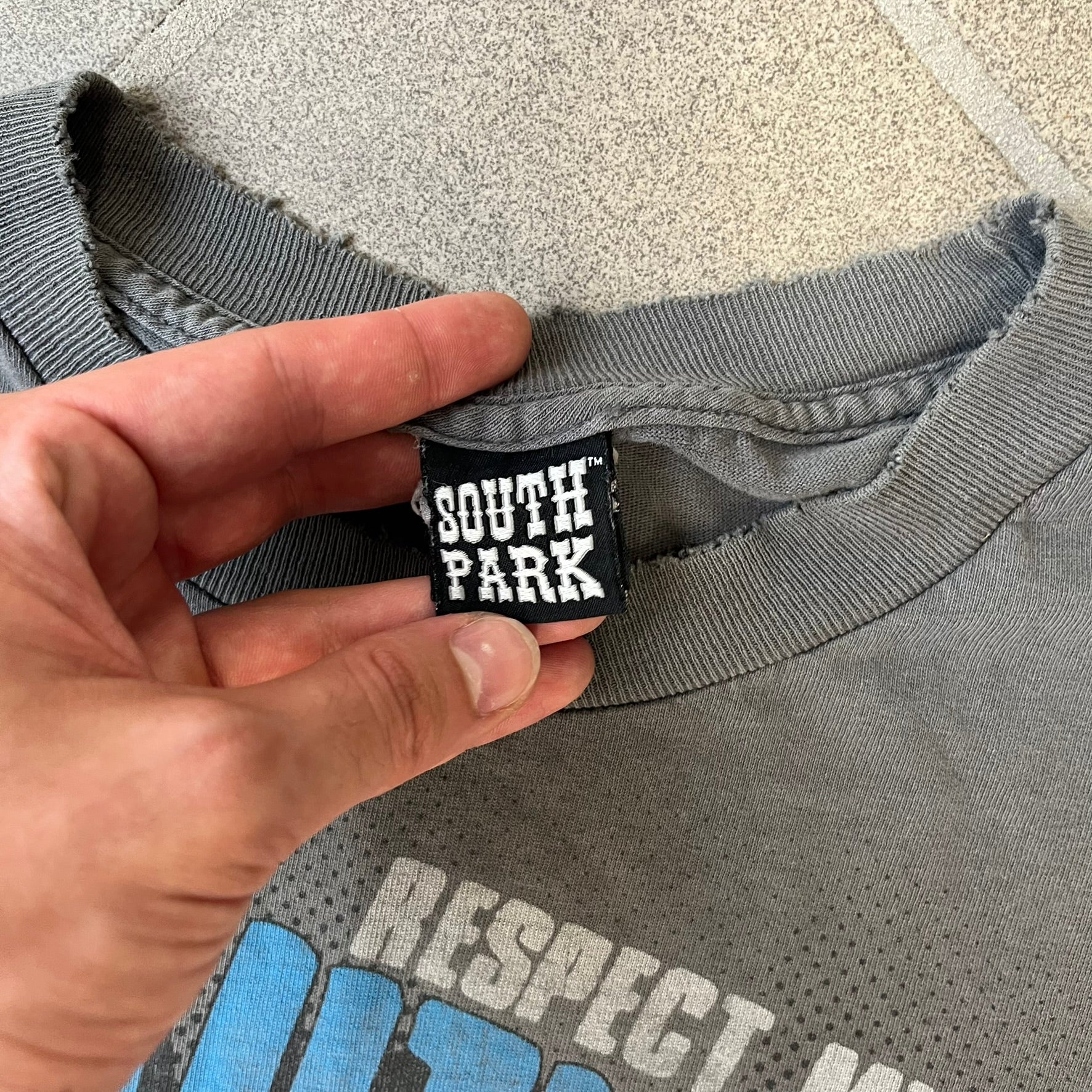 South Park 2009 “Respect My Authority” Gray Tee features a grey T-shirt with a front graphic of Cartman wearing a police uniform and running across a dotted cartoon-style background. Above the graphic is white and blue “Respect My Authority” text. The shirt has a South Park 2009 licensed neck tag and a heavily worn, distressed condition.
