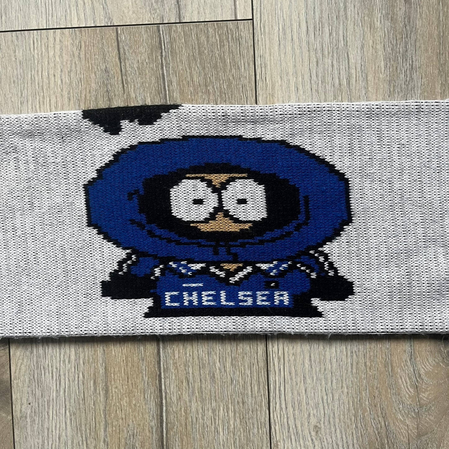 Vintage football scarf in white and black with blue fringe / tassels, featuring South Park characters including Kenny, Cartman, and Kyle dressed in Chelsea kits. The character graphics appear on both sides of the scarf, with consistent coloring in blue. Soft knit construction with a classic football scarf shape and finish.