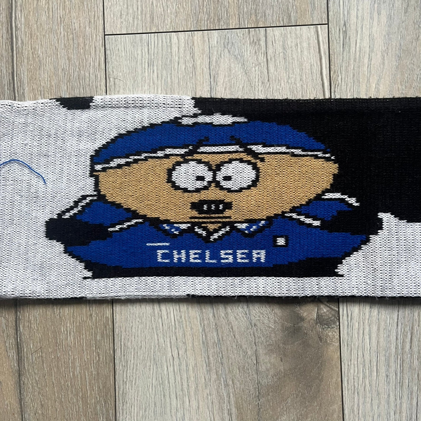 Vintage football scarf in white and black with blue fringe / tassels, featuring South Park characters including Kenny, Cartman, and Kyle dressed in Chelsea kits. The character graphics appear on both sides of the scarf, with consistent coloring in blue. Soft knit construction with a classic football scarf shape and finish.