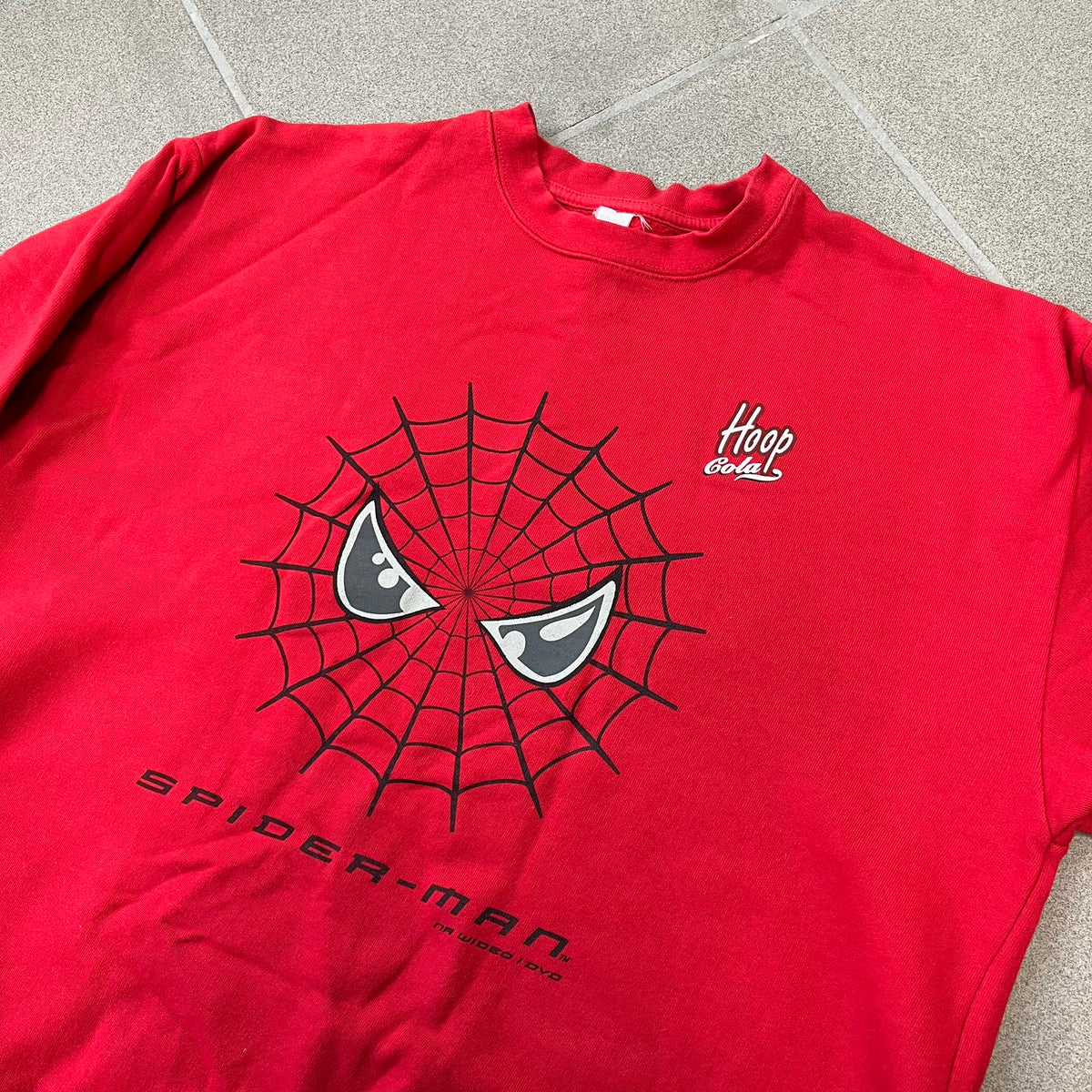 Spider-Man 2002 Hoop Cola Promo Red Crewneck is a rare Poland-exclusive crewneck made for the cross-promotional campaign between Hoop Cola and Warner Bros for the Spider-Man (2002) film. It was never sold in stores and was given out only as a prize in the SMS code contest. The cotton crewneck features web with eyes graphic in the middle, Spider-Man logo under and a Hoop Cola logo above.