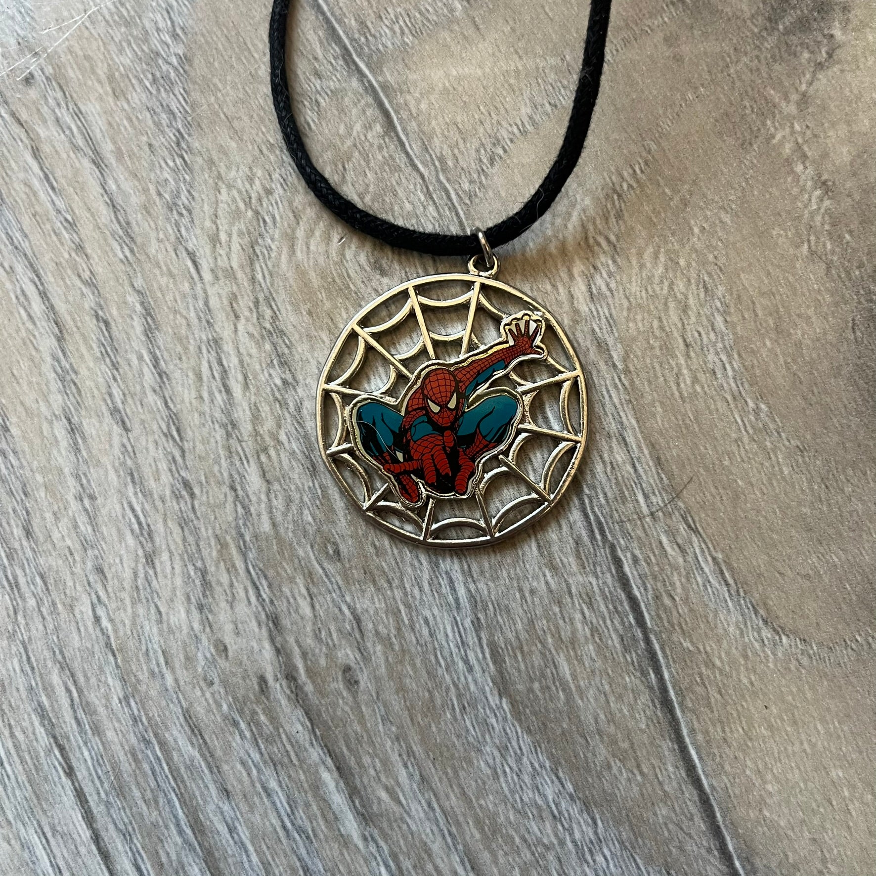 Spider-Man 2006 Web Silver Necklace is a necklace with a black cord and a circular silver-tone metal pendant. The front features a web background with a colored Spider-Man figure in a swinging pose, while the back is plain metal with 2006 licensing.