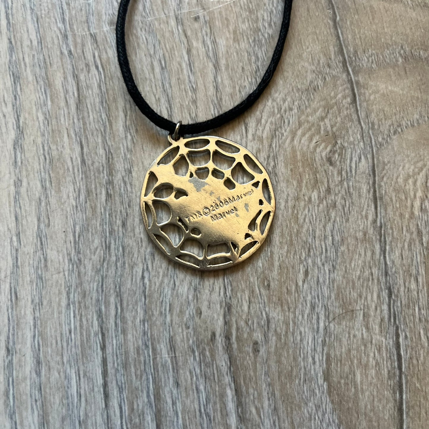 Spider-Man 2006 Web Silver Necklace is a necklace with a black cord and a circular silver-tone metal pendant. The front features a web background with a colored Spider-Man figure in a swinging pose, while the back is plain metal with 2006 licensing.